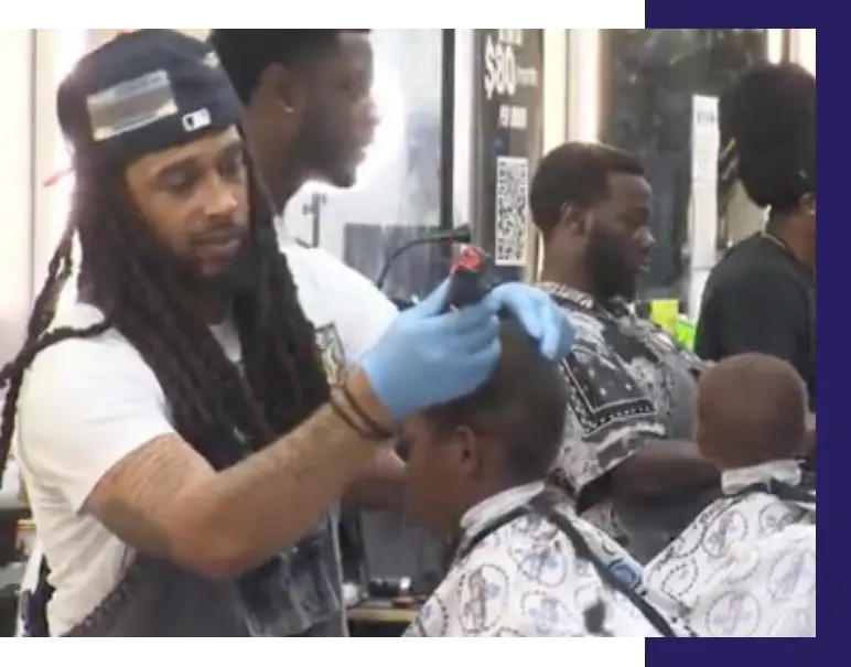A Black barber giving a haircut to a young Black person at a B.A.R.B.E.R.S.H.O.P. event.