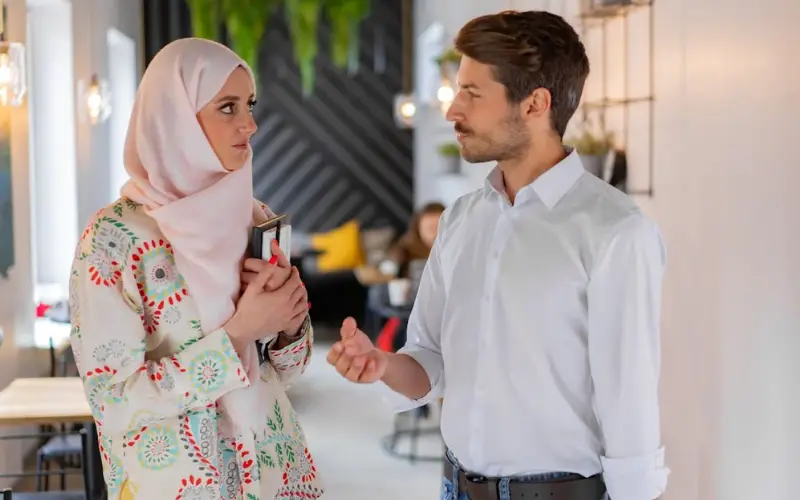 A young, White, male professional talks to a young, Arab, female professional in a hallway.