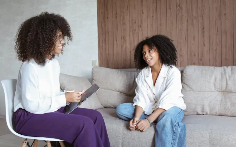 A young Black woman sits on a couch and talks with a young Black mental health professional.
