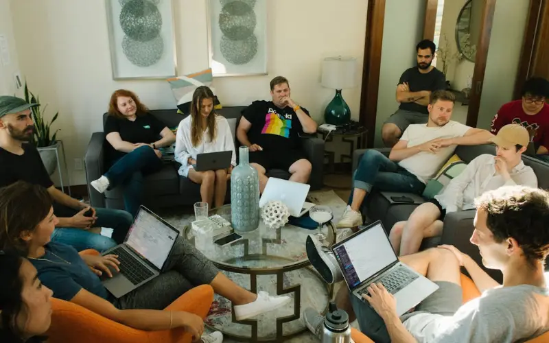 A diverse group of young people sits close together around a coffee table during a large group therapy session.