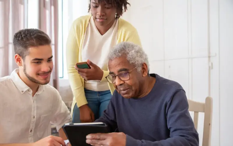 Two young, multiracial people show an older Black male something on a tablet.