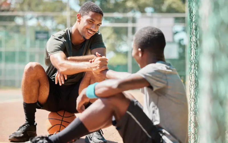 Two young, Black individuals sit on a basketball court and talk.