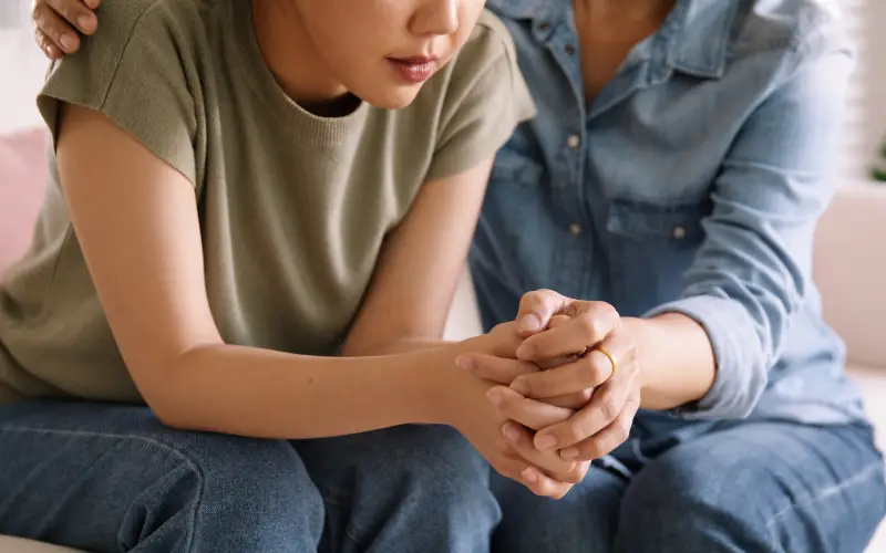 A parent hugs her daughter as she sits on the couch and leans forward.