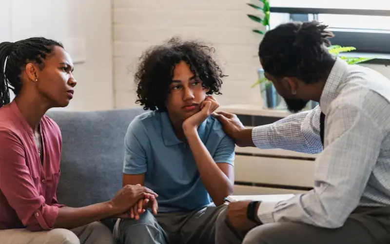 A young, multiracial boy and his mom look focused while they talk to a multiracial, male therapist.